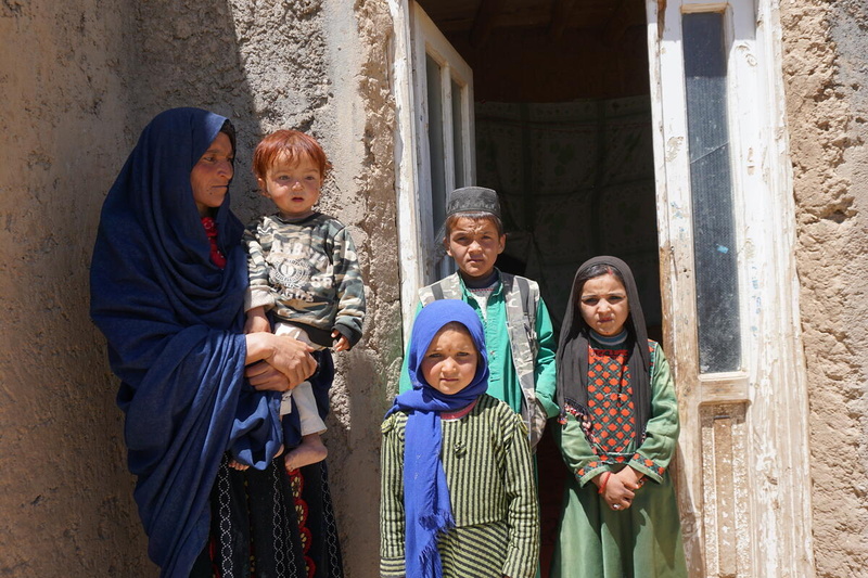 A woman wearing a headscarf and carrying a child stands outside the doorway to a house with three other children