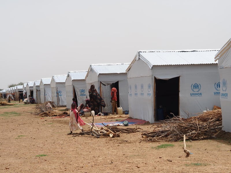 Women and children walk past a row of UNHCR-branded shelters.