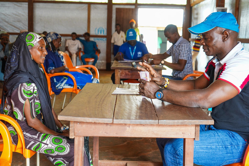 A woman sits across a desk from a man in a blue UNHCR cap as he counts out money.