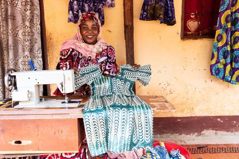 A smiling woman sitting next to a sewing machine table holds up a green and white patterned dress