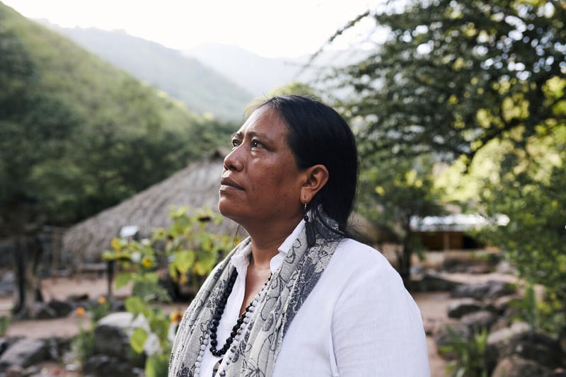 A woman in a white shirt stands in the middle of a village with trees and mountains in the background.