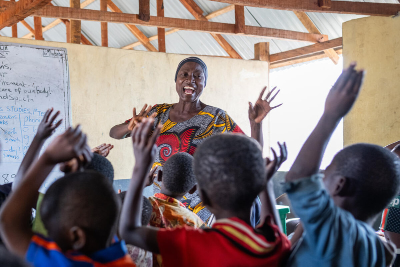 A woman leads a class of young children with their hands in the air.