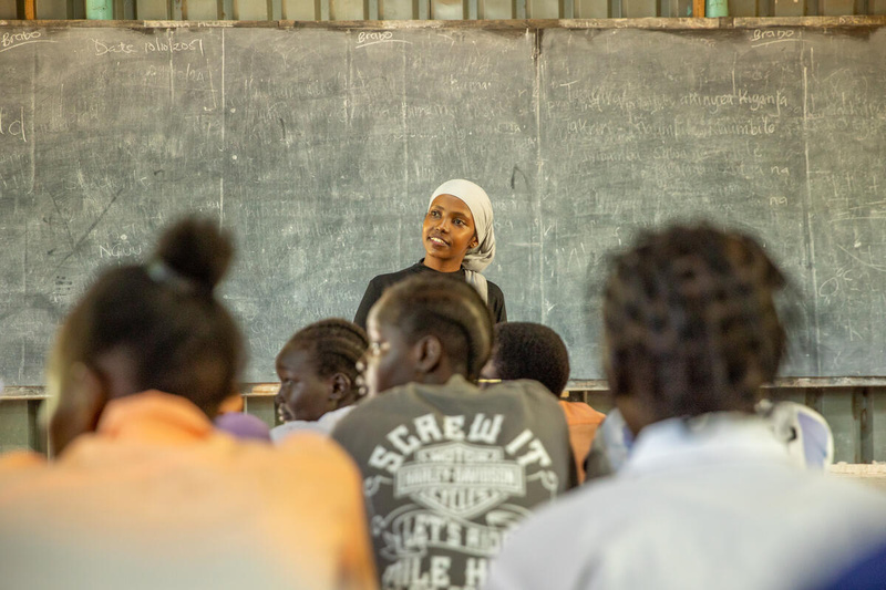 A young woman stands in front of a blackboard addressing a class of other young women.