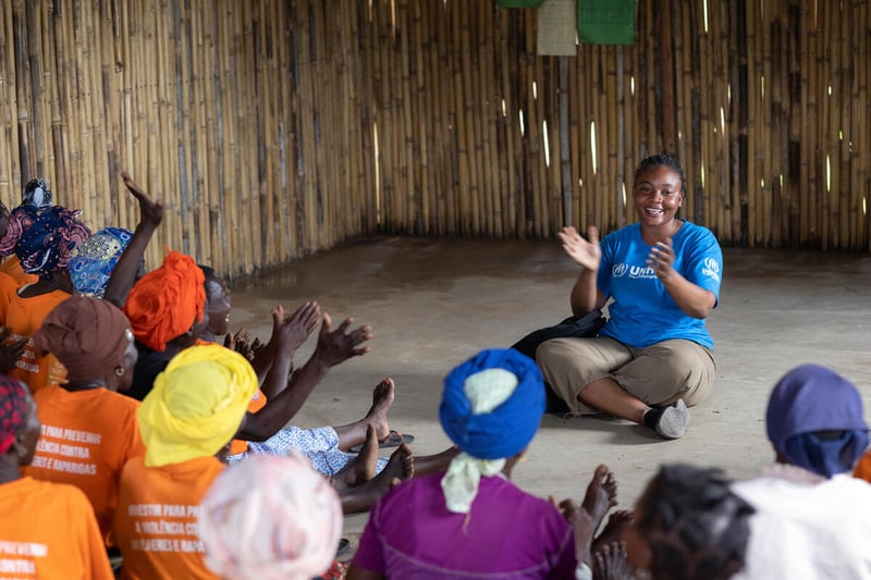 A woman in a UNHCR branded t-shirt sits on the floor leading a group of women in clapping.