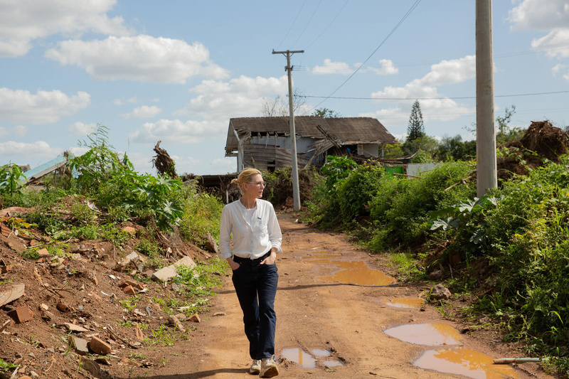Cate Blanchett walks along a dirt road with puddles. Around her is destruction caused by flooding – behind her, a damaged building and on the sides of the road, building debris.