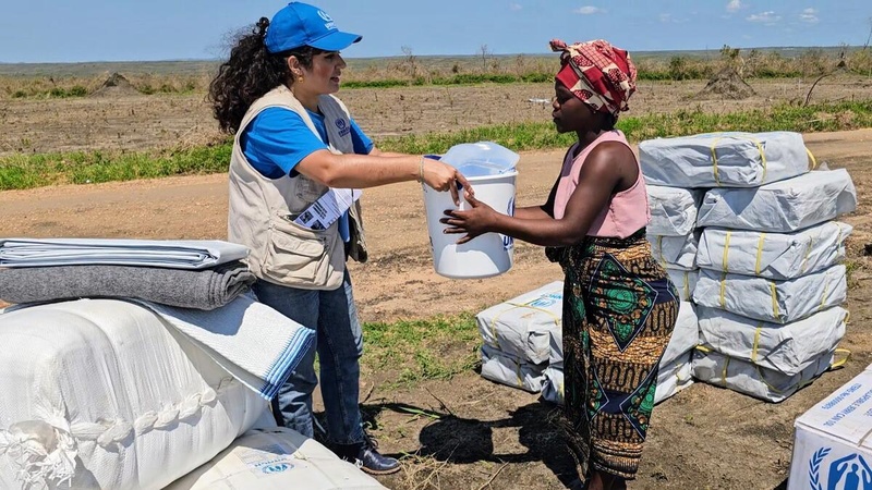 A woman in a UNHCR-branded vest and hat hands a bucket of relief items to a woman.