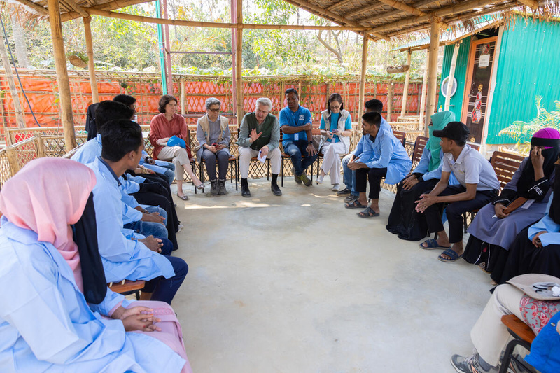 A man addresses a group of people seated in an open-sided shelter.