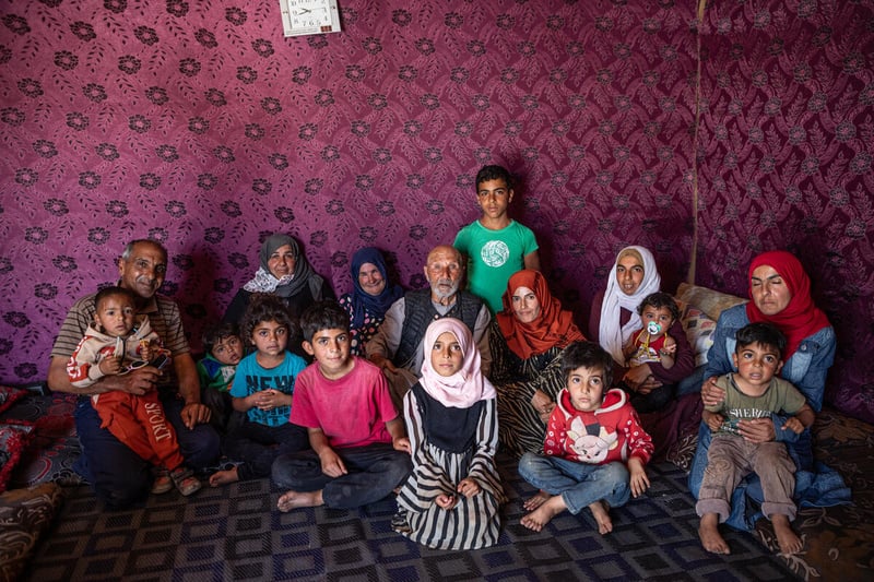 A large family of several generations sits on the carpeted floor inside a shelter