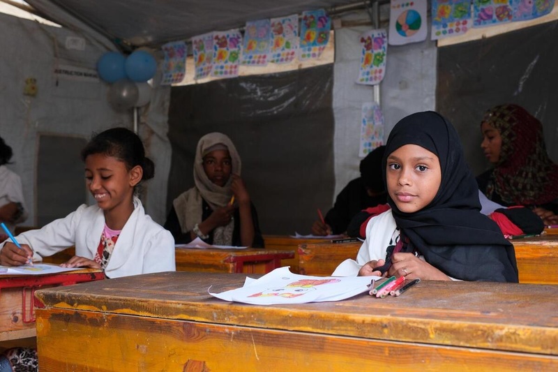 Girls sit at wooden desks with paper and coloured pencils in classroom inside a tent.