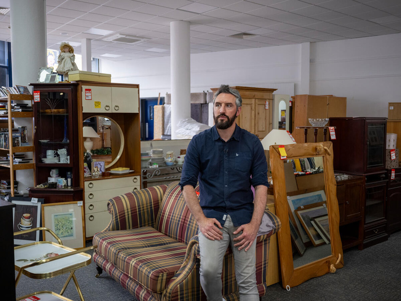 A man wearing a blue shirt and jeans leans on the arm of a sofa in a second-hand furniture store.