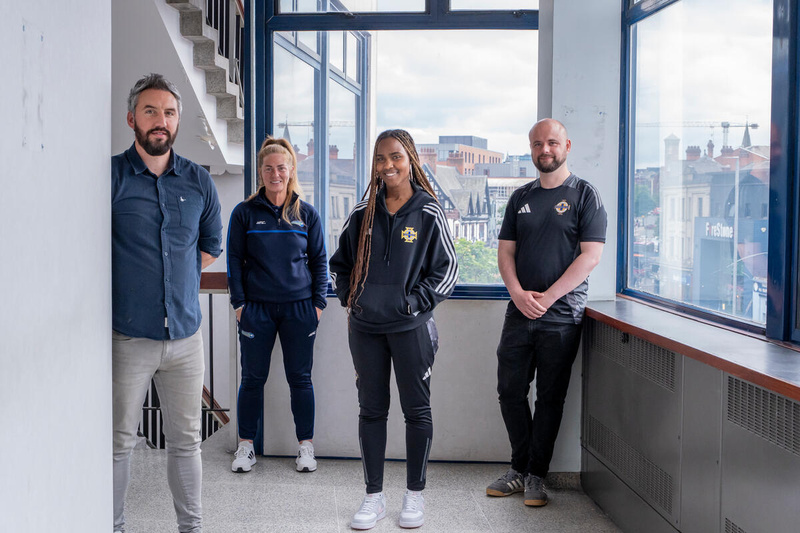 A group of four people stand on the landing of a building in front of large windows overlooking a city.