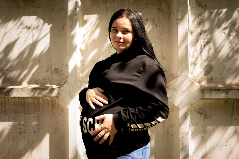 A pregnant woman with dark hair wearing a back pullover cradles her stomach as she stands outside in front of a sun-dappled wall