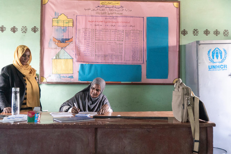 A woman sits behind a teacher's desk writing at the front of a classroom as another woman stands on her right
