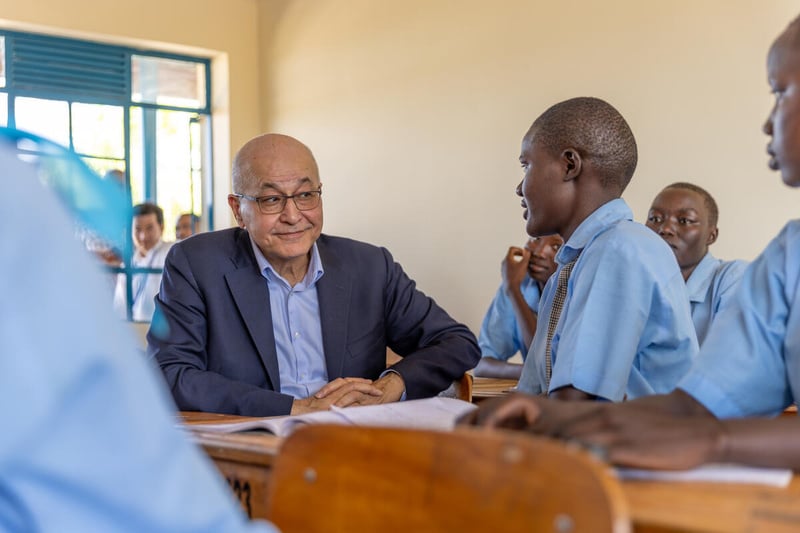 A man in a suit talks to a pupil in uniform as they sit at school desks in a classroom full of students