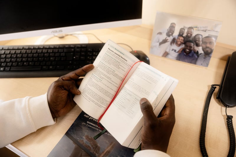 A man's hands hold an open book above a desk holding a computer, a telephone and a photo of a group of friends