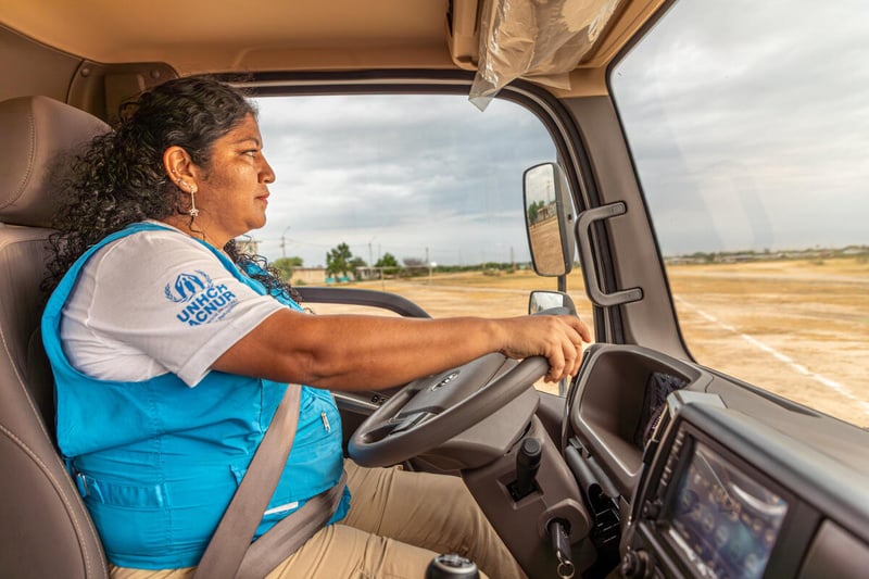 A woman wearing a blue UNHCR vest drives a truck across a dusty soccer field.