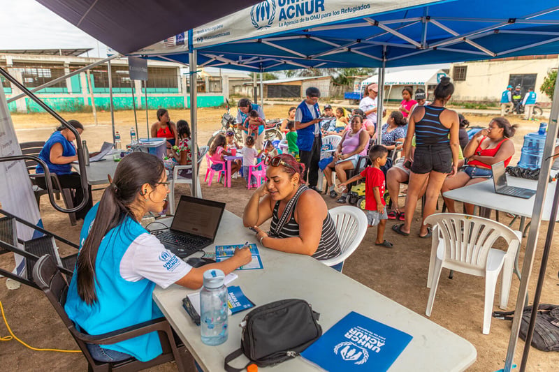 Women and children sit on chairs under a large shaded awning accompanied by UNHCR staff 