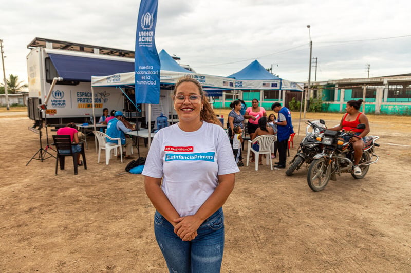A woman in a white t-shirt and blue jeans stands in front of a group of people with a truck in the background