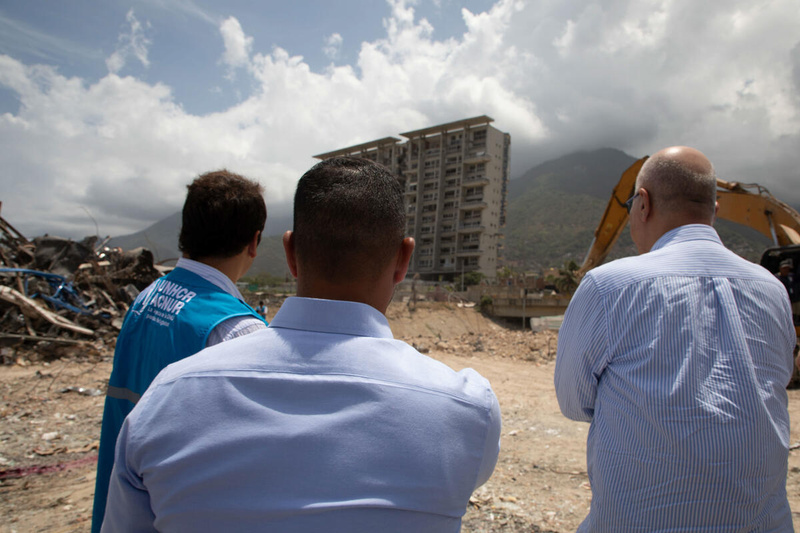 Three men stand outside looking towards a tower block surrounded by mounds of rubble with a mechanical digger in the background