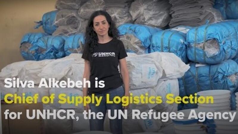 A UNHCR staff member stands in front of supplies Text on image reads ''Silva Alkebeh is Cheif of Supply Logistics Section for UNHCR, the UN Refugee Agency'.