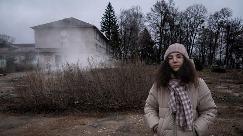 A girl stands in front of a damaged school building.