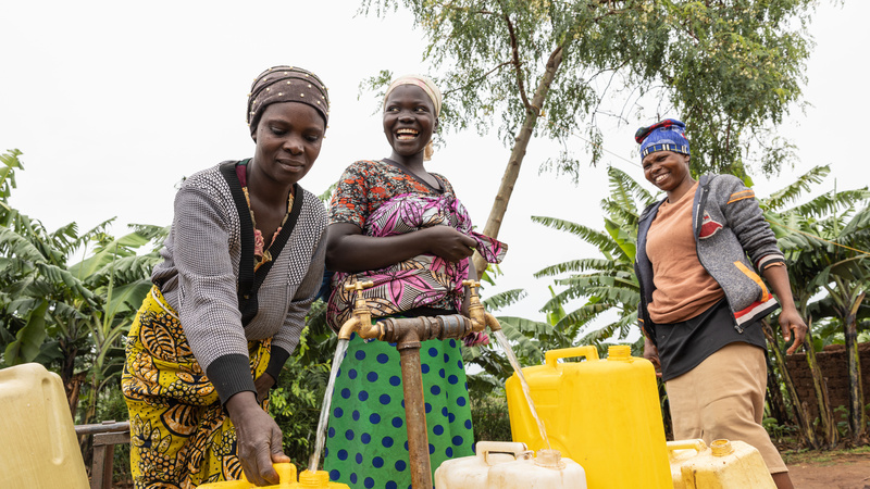 A woman fills up a container at a tap while two other women watch on.