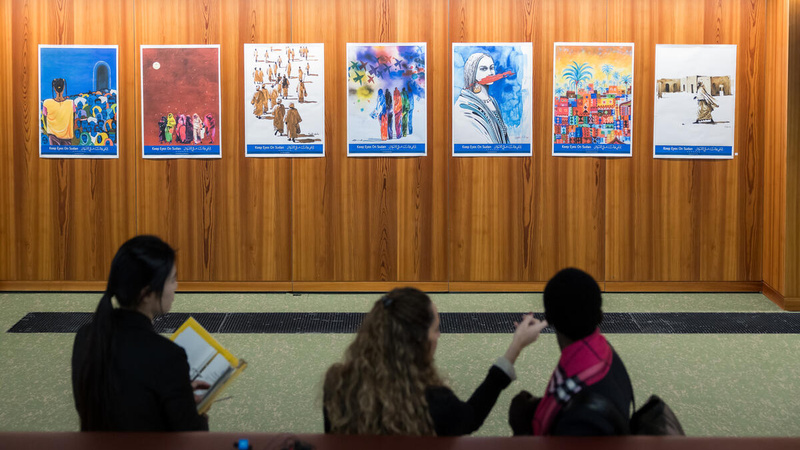 Three persons sit on a couch, looking at several artworks displayed on a wall in front of them.