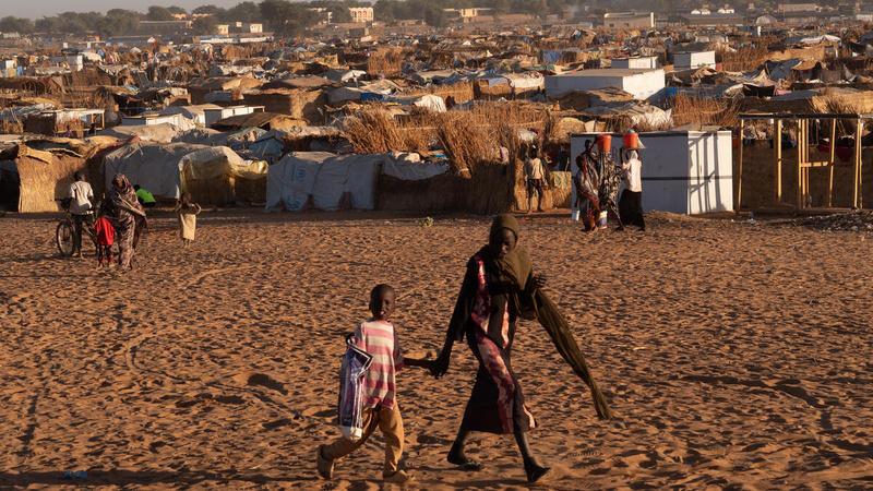 A woman holds a child's hand as they walk past a refugee tent settlement in a dusty field.