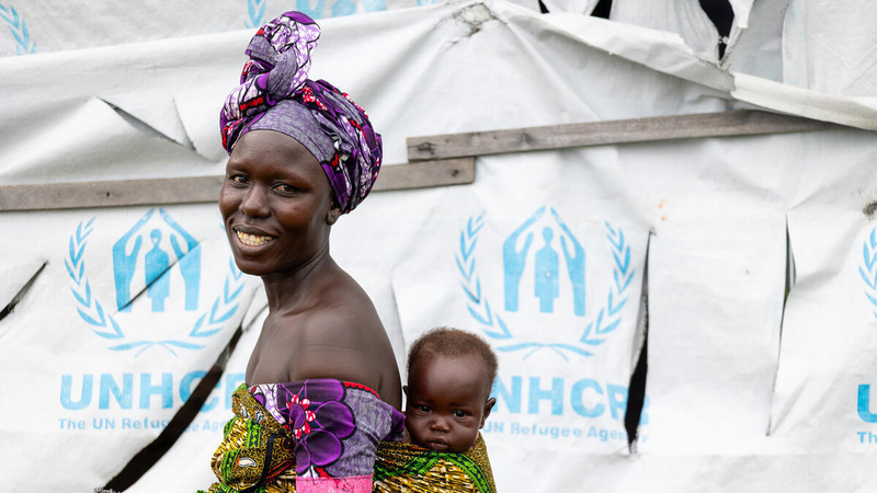 A smiling woman carries her baby boy on her back in front of a UNHCR tent.