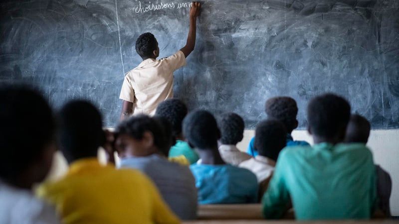 A man writes on a blackboard in a classroom of teenagers. 