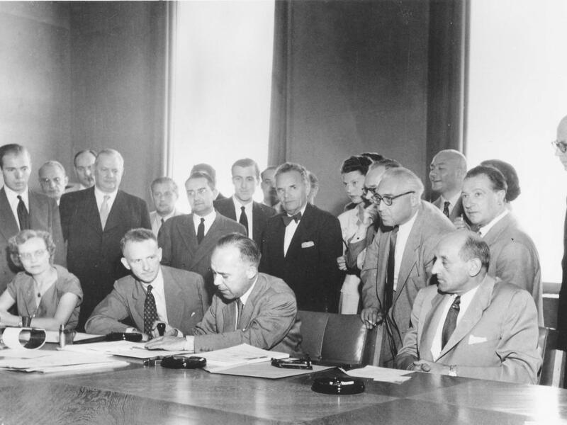 Back and white photo: A man signs the 1951 Refugee Convention, surrounded by a group of people.