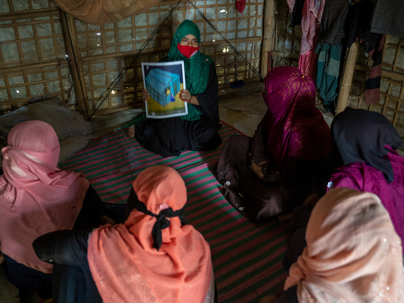 A woman seated in a bamboo shelter holds up an educational poster for other women to see who are seated around her.