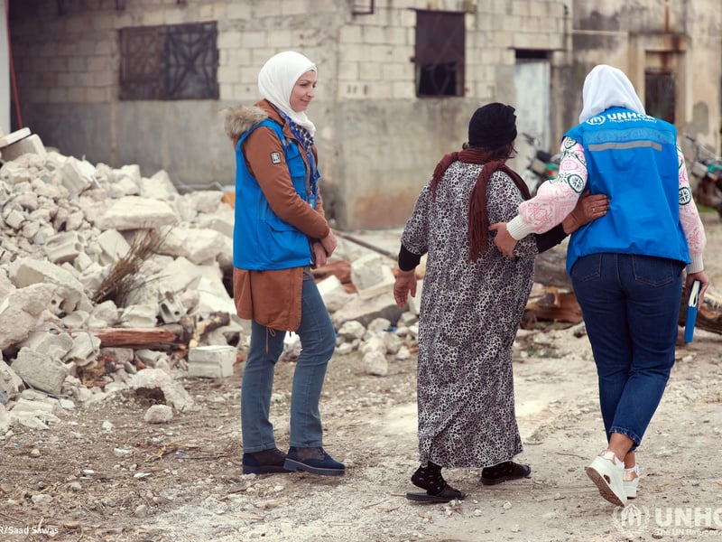 Deux employées du HCR marchent avec une personne déplacée syrienne à côté des débris de sa maison.