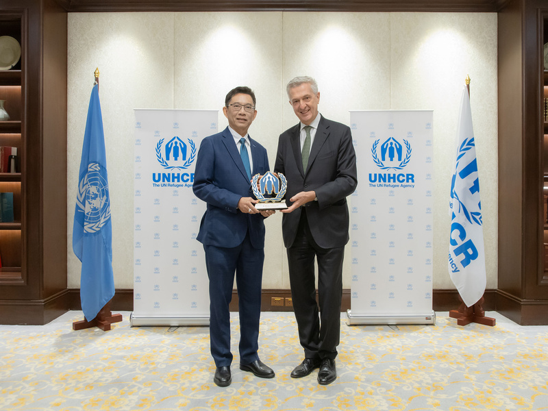 Mr. Wanchai Tachavejnukul and Filippo Grandi smiling and holding an award together, standing in front of UNHCR banners and flags