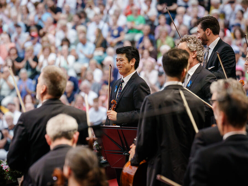 Musicians part of an orchestra stand up holding their instruments, in front of a crowd.
