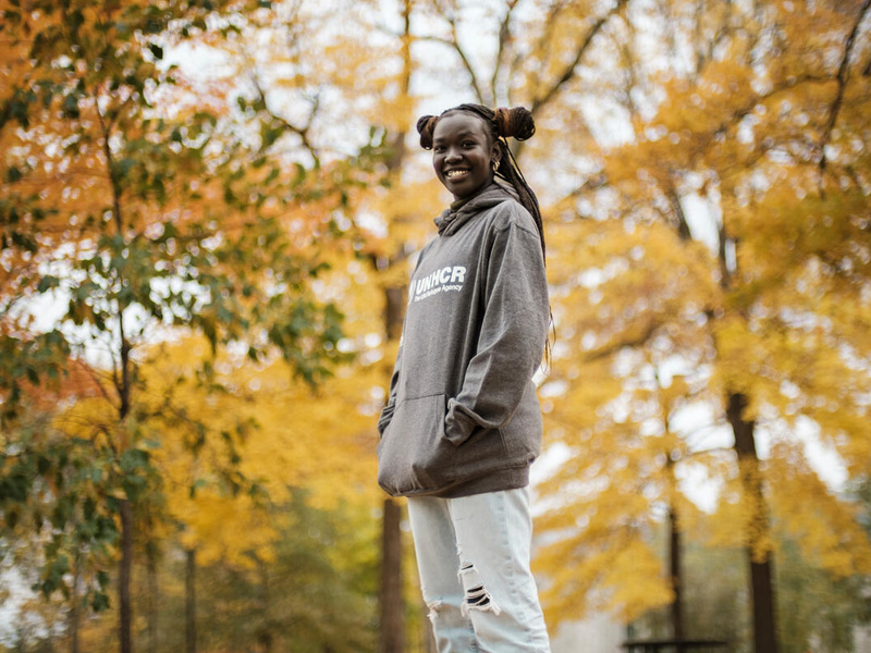Mary Maker stands in a garden, below trees with autumn-coloured leaves.