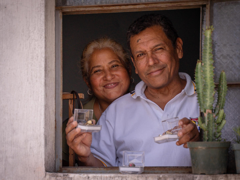 A man and woman, framed in a shop window, smile and hold up items of handmade jewellery.