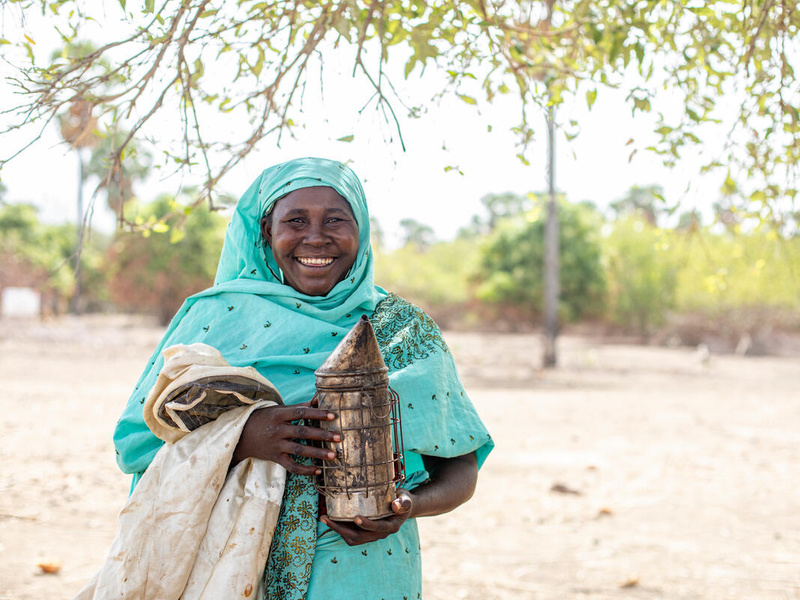 A woman in a colorful headscarf holds beekeeping equipment and smiles for the camera.