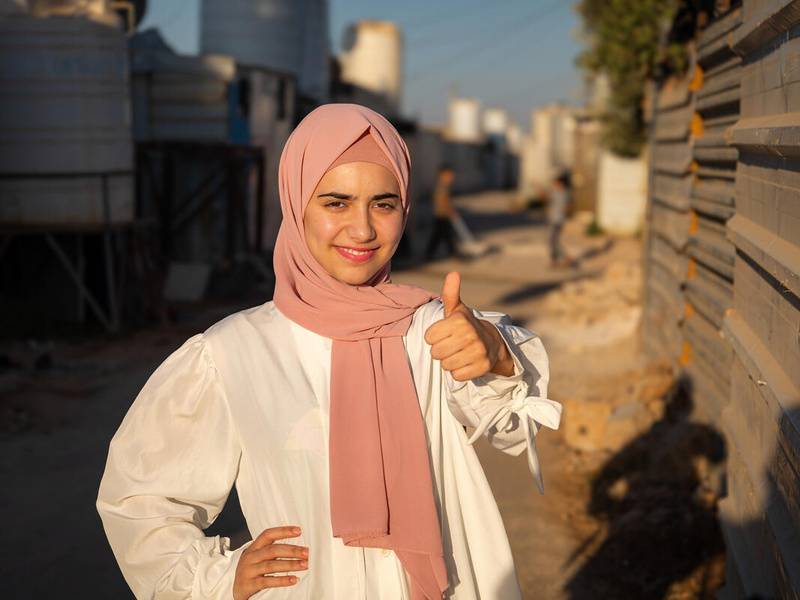 A young woman in a pink headscarf smiles confidently and gives the camera a thumbs up.