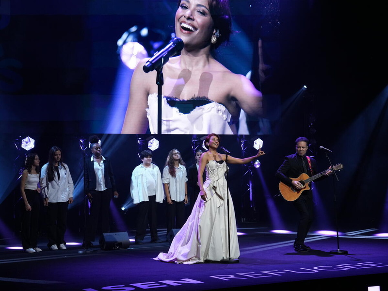 A woman performs onstage with a guitarist and backing singers in front of a giant screen