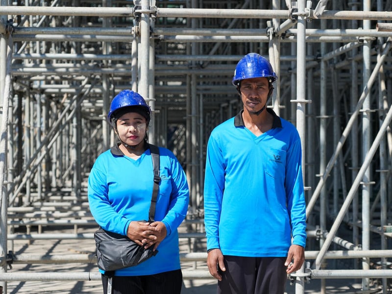 A woman and a man wearing matching blue shirts and hard hats stand outside in front of scaffolding