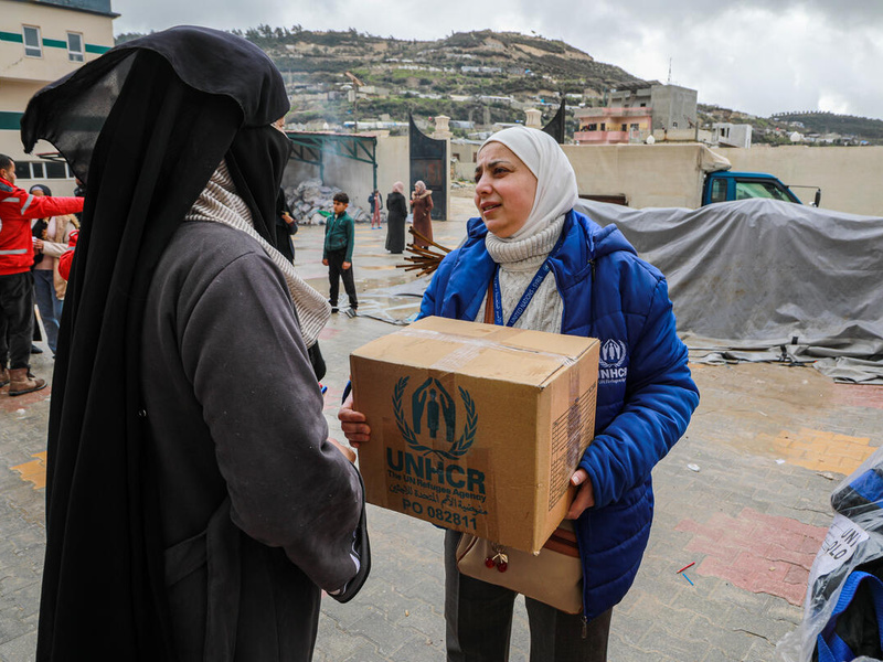 A woman in a UNHCR jacket hands a box of UNHCR supplies to another woman