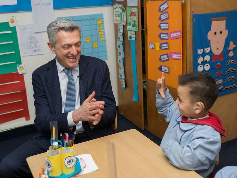 UNHCR High Commissioner Filippo Grandi sits with a 7-year old Syrian refugee child in a classroom. The child is wearing a hearing aid.