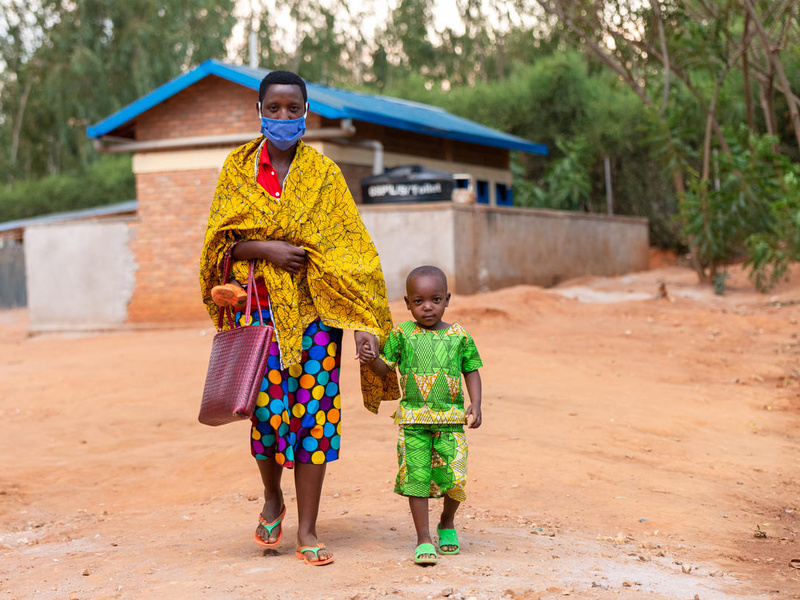 A Burundian woman wearing colourful clothes and a mask hold her child's hand.