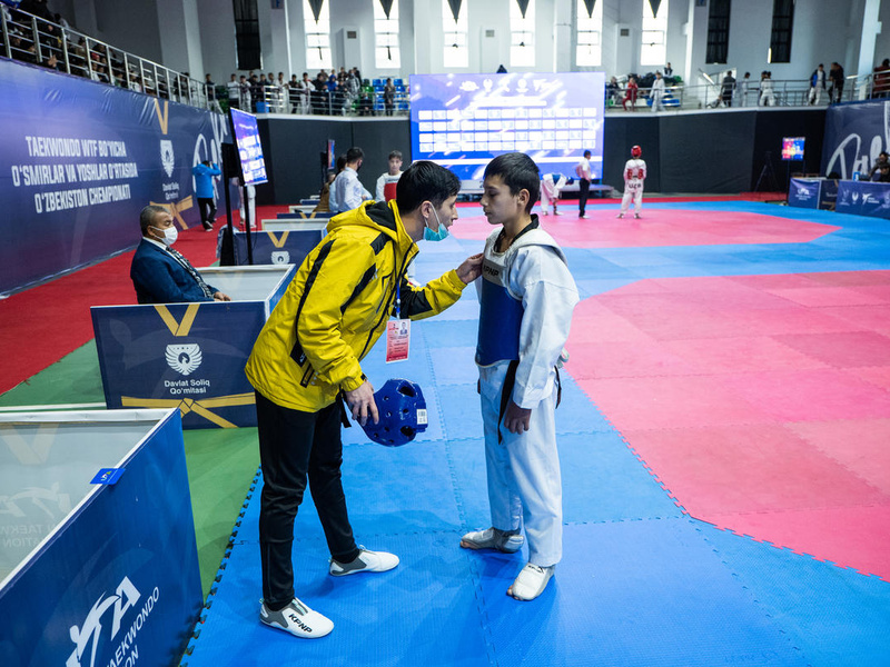 A man in a bright yellow athletics tracksuit speaks to a young child in a martial arts uniform inside a gymnasium.