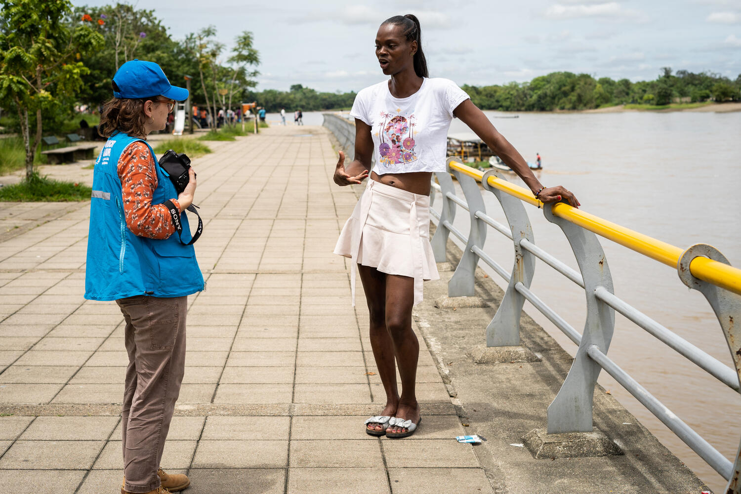 A woman in a UNHCR-branded vest talks to another woman as they stand next to a river.