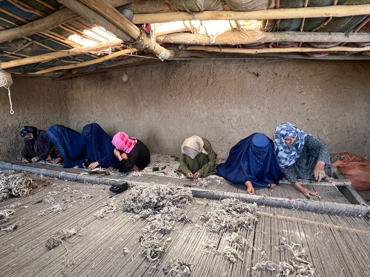 Seven women are bent over a giant weaving loom on the floor of a mud-brick room with a large holes in the roof.