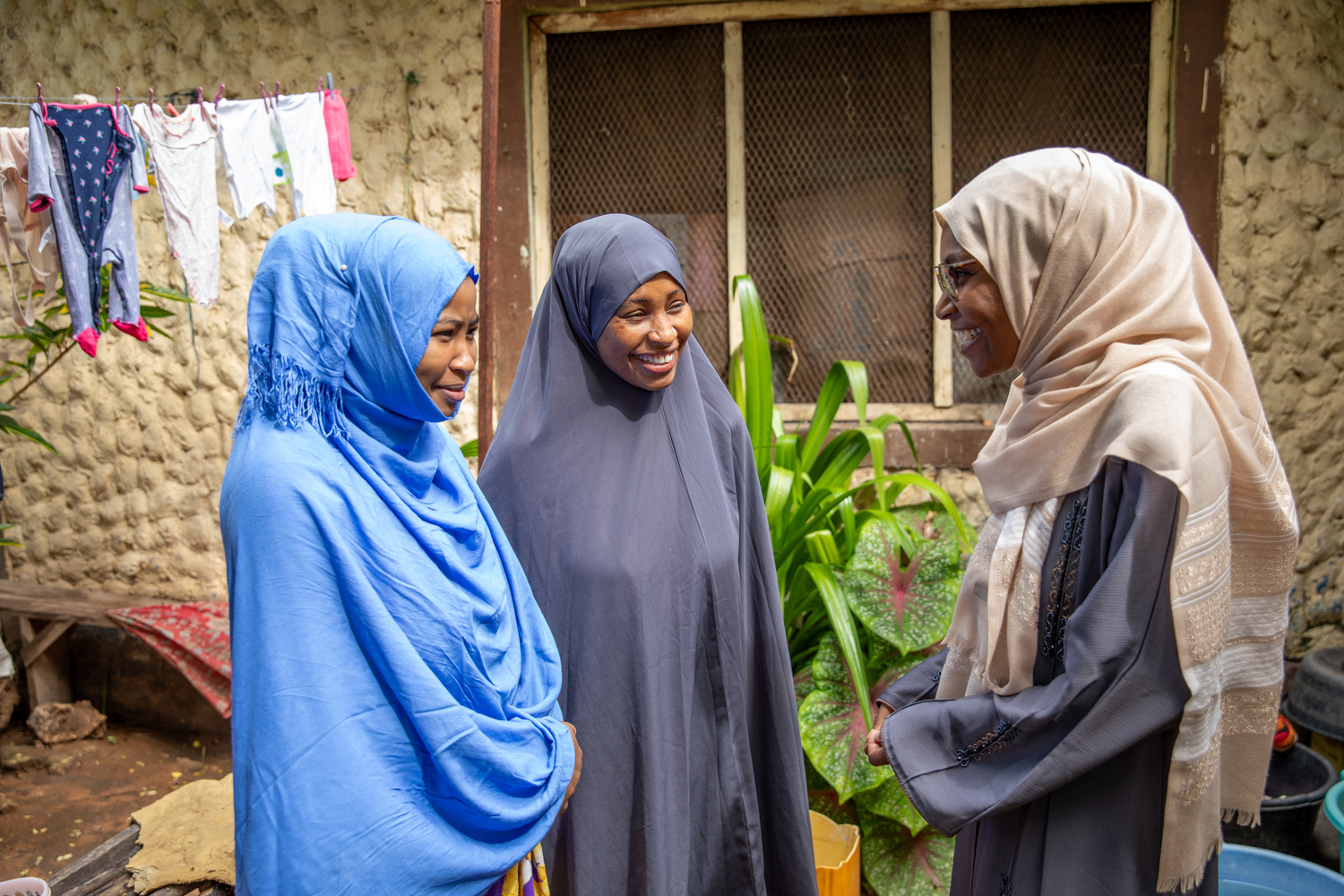 Three smiling women stand together in front of a house.