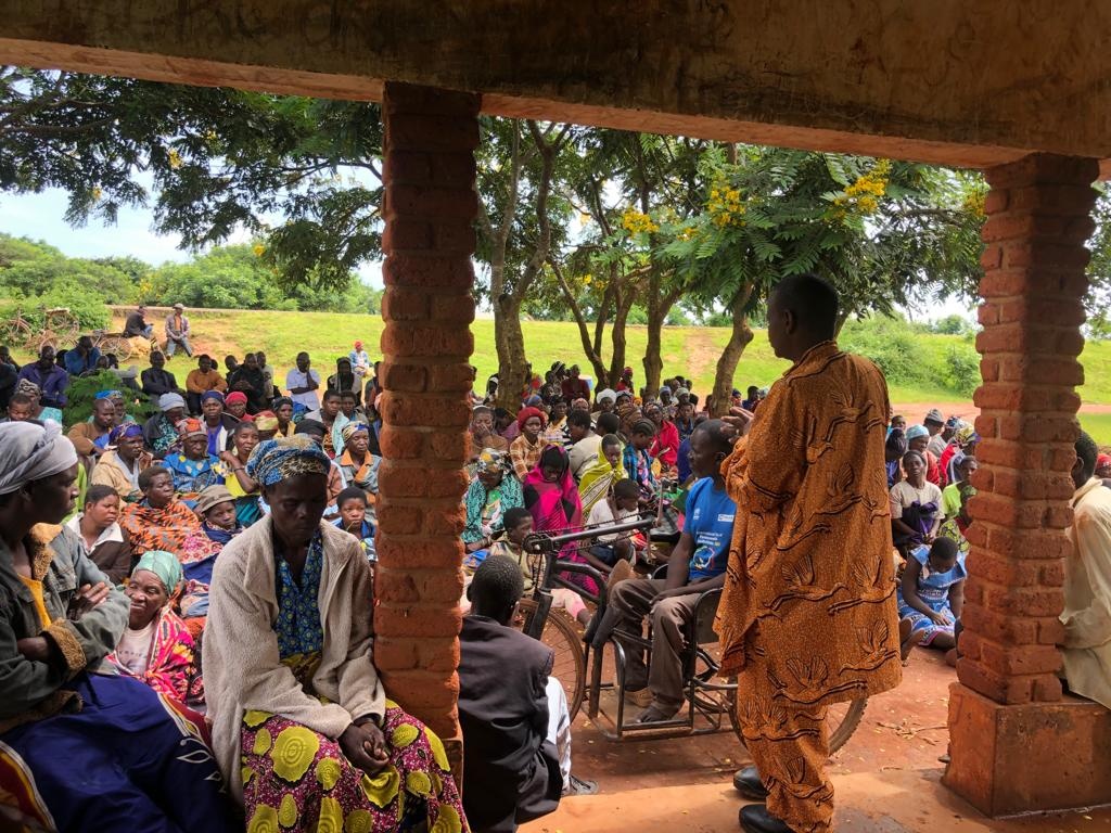 A large group of colourfully-dressed people celebrate International Disability Day outside under trees, pictured from under a brick shelter