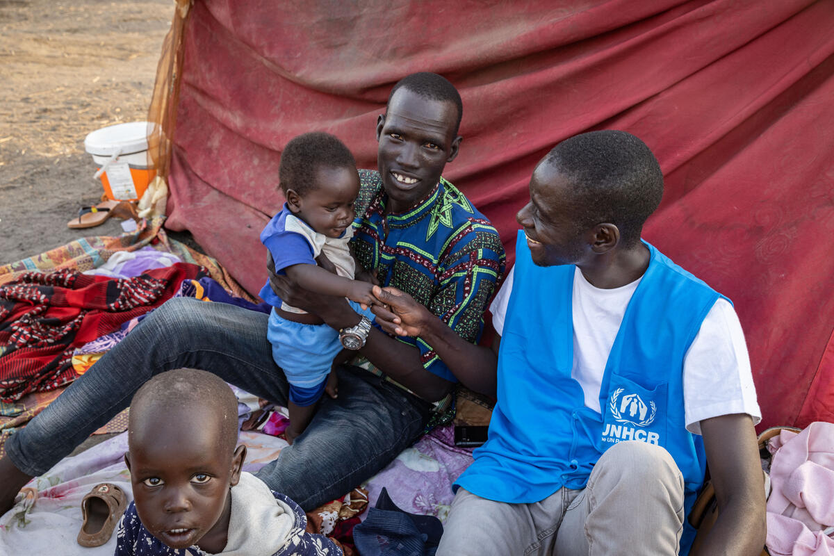 Two men, a returnee and a UNHCR staff member, sit together on a blanket on the ground with a baby and a small child.
